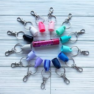 May include: A variety of colorful silicone keychains with silver key rings and clips. The keychains are shaped like small bottles and are arranged in a circle around a pink lip balm tube with the text "Lip Smacker" and "Coconut" on the label.