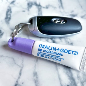 May include: A small, white tube of Malin+Goetz lip moisturizer with a purple cap. The tube is labeled with the brand name, product name, and size: 0.3 fl. oz. e 10 ml.
