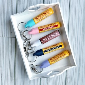 May include: Five Burt's Bees lip balms with keychains attached, arranged in a white tray. The lip balms are in the flavors: Beeswax, Strawberry, Tinted, Vanilla Bean, and Beeswax.