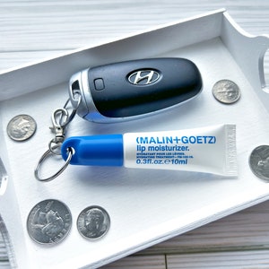 May include: A white tray with a blue and white lip balm tube, a silver key chain with a car key, and several silver coins. The lip balm tube has the text "(MALIN+GOETZ) lip moisturizer. HYDRATANT POUR LES LEVRES. HYDRATING TREATMENT-P-100 0.3fl.oz.e10ml" printed on it.