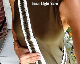 May include: A crochet crossbody strap featuring a dark olive green center stripe bordered by white edges. The strap includes a silver-toned metal adjustable slider and a swivel clasp attached to a black phone case. The text Inner Light Yarn is visible.