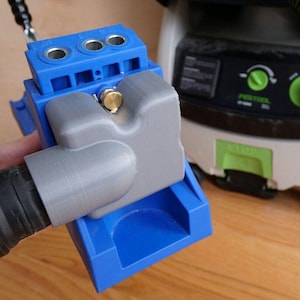 May include: A blue and gray woodworking jig with a dust port for connecting to a vacuum. The jig has three drill bit holes and a depth stop.