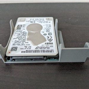 Può includere: Un alloggiamento per hard disk grigio con un hard disk argentato all'interno. L'hard disk ha il testo "HGST" e "www.hgst.com" stampato su di esso.