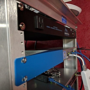 Könnte beinhalten: Ein Serverschrank mit verschiedenen elektronischen Komponenten. Der Schrank verfügt über eine schwarze Steckdosenleiste, einen blauen Netzwerk-Switch und mehrere Kabel. Die Komponenten sind in einem Metallrahmen montiert, mit einer roten Wand im Hintergrund.