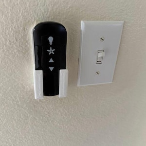 May include: A white wall mounted light switch with a black remote control for a ceiling fan. The remote control has buttons for light, fan speed, and direction.