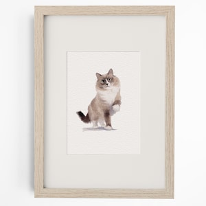 May include: A watercolour painting of a fluffy cat with brown and white fur, depicted in a sitting position with one paw raised, presented inside a light-coloured wooden frame with a white mat board border surrounding the artwork.