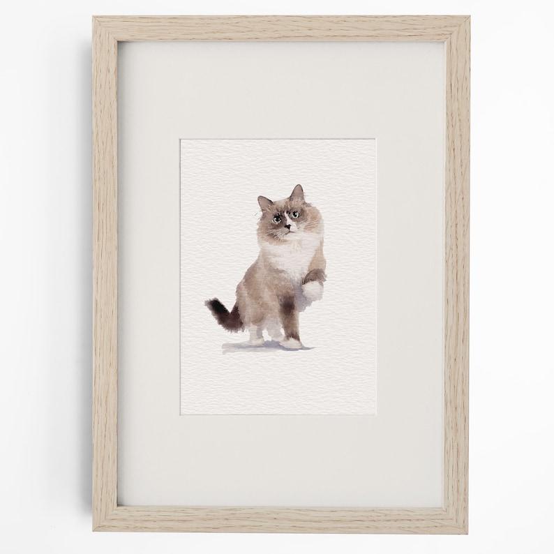 May include: A watercolour painting of a fluffy cat with brown and white fur, depicted in a sitting position with one paw raised, presented inside a light-coloured wooden frame with a white mat board border surrounding the artwork.