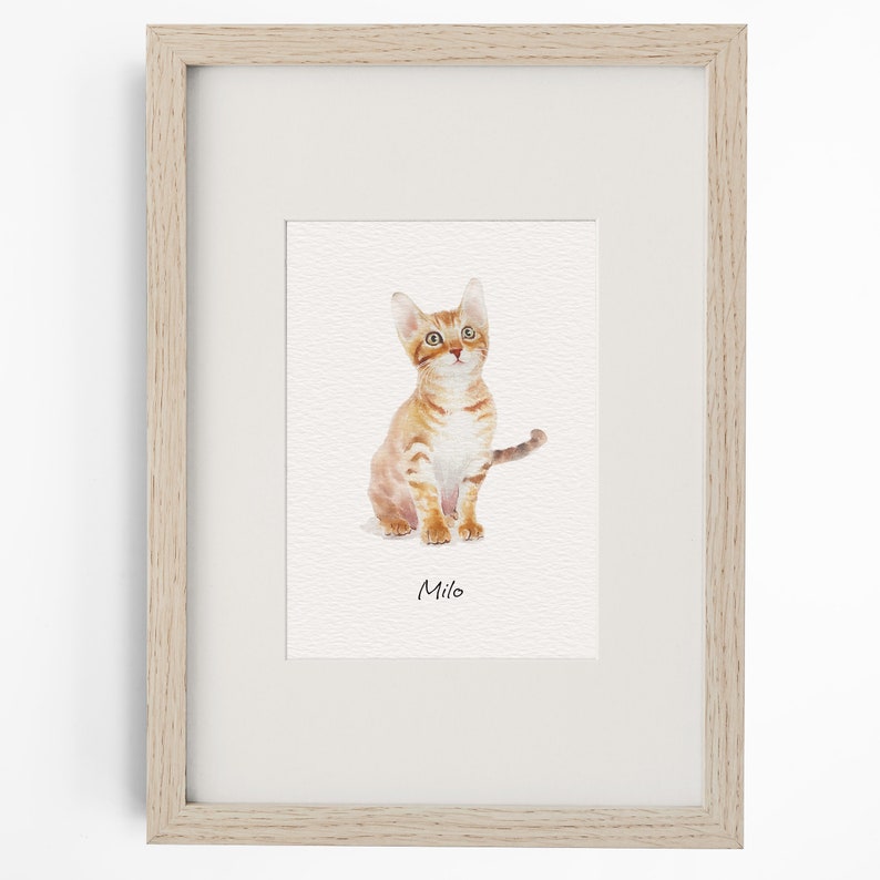 May include: A framed watercolour print featuring an orange tabby kitten sitting and looking upward. The kitten has light orange fur with darker stripes and green eyes. Below the kitten, the name Milo is printed in a simple black sans-serif font.