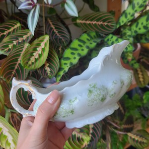May include: White porcelain gravy boat with a green floral design. The boat has a scalloped edge and a gold-trimmed handle.