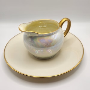 Puede incluir: Una jarra de crema blanca con un borde dorado y un acabado iridiscente se asienta sobre un platillo a juego con un borde dorado.