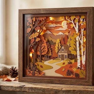 May include: A square wooden shadow box frame containing a multi-layered paper cut art scene of an autumn forest landscape. The scene features a small house, a winding path, birch trees, and mountains under a crescent moon, illuminated by warm internal lights.