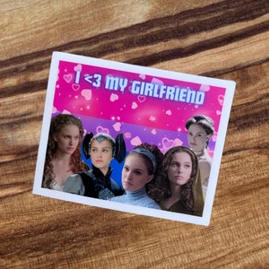 May include: A rectangular sticker with a pink and blue background featuring images of women and the text "I <3 MY GIRLFRIEND" in white. The sticker has a white border and is placed on a wooden surface.