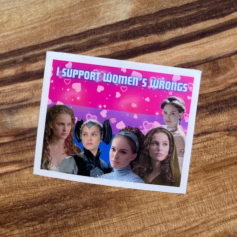 May include: A rectangular sticker with a pink and blue background, featuring images of women and the text "I SUPPORT WOMEN'S WRONGS." The sticker has a white border and is placed on a wooden surface.