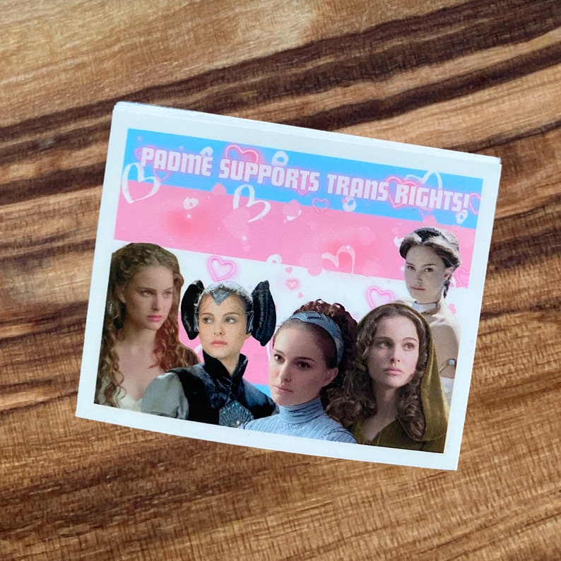 May include: A white sticker featuring images of Padm&eacute; Amidala from Star Wars. The sticker has a pink and blue background with hearts and the text "Padm&eacute; Supports Trans Rights!"