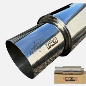 Puede incluir: Un silenciador de escape HKS cromado pulido con una punta inclinada. El silenciador tiene el logotipo y el texto de HKS. Una versión más pequeña del silenciador se muestra en una caja de cartón con el logotipo de HKS.