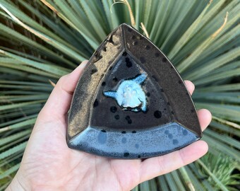 May include: A triangular ceramic dish with a dark brown glaze and a light blue fish design in the center. The dish has a speckled texture and a blue rim. The dish is held in a hand.