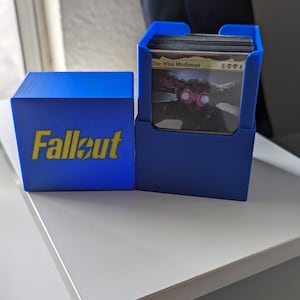 May include: A blue card storage box with the word "Fallout" printed on the front. The box is open and a card with the text "The Wise Mothman" is visible.