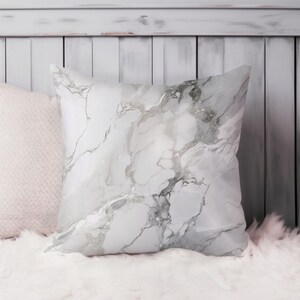 Peut inclure: Coussin carré décoratif avec un motif de marbre blanc et gris. Le motif présente des veines grises fluides. Le coussin est posé sur une surface blanche et duveteuse, avec un aspect doux et texturé.