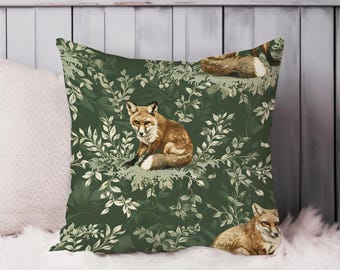 May include: Square decorative pillow with a forest theme. The pillow features a dark green background with a repeating pattern of leaves and several foxes in shades of brown and white. The pillow is placed on a white fluffy surface.