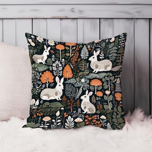May include: Square decorative pillow with a dark background featuring a repeating pattern of white rabbits, orange and white flowers, and green foliage. The pillow is placed on a white fluffy surface.