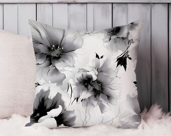 May include: Square decorative pillow featuring a large floral pattern in shades of gray and black on a white background. The pillow has a soft, fluffy texture and is placed on a white surface. The floral design includes detailed petals and leaves.