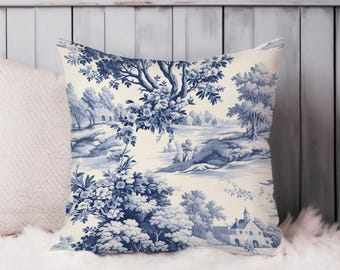 May include: A square throw pillow with a blue and white toile pattern. The design features a pastoral landscape with trees, buildings, and floral elements. The pillow is on a white surface.