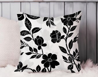 May include: Square throw pillow with a black floral pattern on a white background. The design features stylized black flowers and leaves. The pillow is placed on a white, fluffy surface.