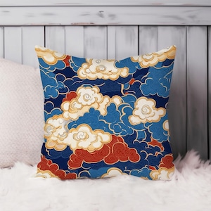 May include: Square decorative pillow with a navy blue background and a repeating pattern of stylized clouds in white, red, and blue. The clouds have gold outlines, creating a striking contrast. The pillow is placed on a white, fluffy surface.