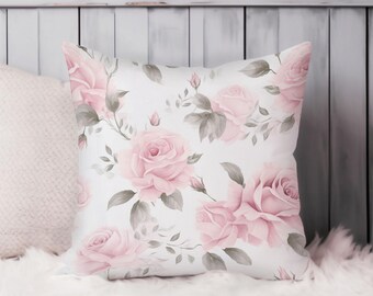 May include: Square throw pillow with a white background and a pattern of large pink roses and gray leaves. The pillow is resting on a white fluffy surface, with a white wooden background.