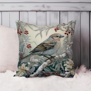 May include: Square decorative pillow featuring a detailed illustration of a bird perched on a snow-covered branch. The bird has brown and gray feathers, with red berries and white flowers in the background. The pillow is on a white surface.