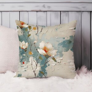 May include: Square cushion with a floral design. The cushion features large white flowers with orange centres and red accents against a textured, light blue and cream-coloured background. The cushion is placed on a white, fluffy surface.