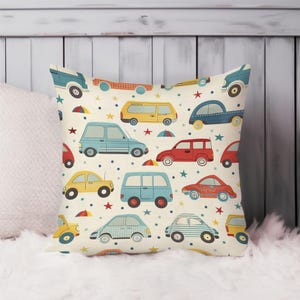 May include: A cream-colored pillow with a colorful pattern of vintage cars, vans, and buses. The pattern includes red, blue, yellow, and white cars with black outlines. The background is a cream color with small white dots.