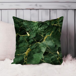 May include: Square decorative pillow with a dark green marble design and gold veining. The pillow is on a white surface with a white wooden background.
