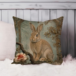 May include: Square decorative pillow featuring a detailed illustration of a brown rabbit perched on a branch with a pink and cream flower. The background is a soft blue with ornate gold-colored framing.