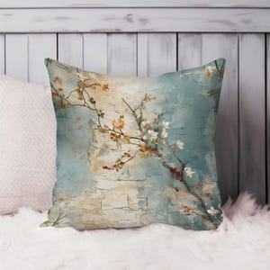 May include: Square cushion with a floral branch design. The cushion has a light blue and beige background with a textured, painted effect. The branch has white and orange blossoms.