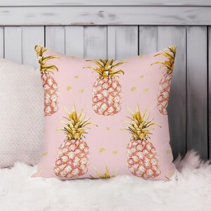 May include: Square decorative pillow with a pink background featuring a repeating pattern of gold and pink pineapples. The pineapples have gold crowns and pink bodies. The pillow is on a white surface.