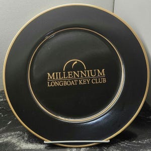 Pode incluir: Um prato preto com borda dourada e círculo interno dourado. O prato tem as palavras "MILLENNIUM LONGBOAT KEY CLUB" em letras douradas. O prato está em um suporte de metal branco.