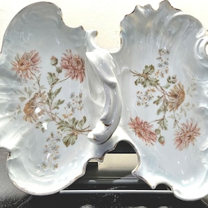 May include: A white, two-sectioned serving dish with a handle, featuring a floral design in shades of pink, brown, and green. The edges are scalloped and trimmed with gold. The dish is likely made of porcelain.