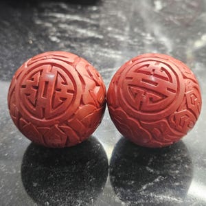 May include: Two red carved spheres with intricate patterns and symbols. The spheres have a detailed, textured surface with a matte finish. The carvings appear to be of a traditional design, possibly Asian-inspired, and are symmetrical.