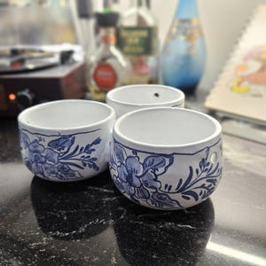 May include: Three white ceramic bowls with a blue floral design. The bowls are round with a slightly flared rim. The floral pattern is hand-painted in a detailed, stylized design. The bowls are on a dark, reflective surface.