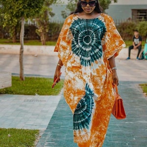 Peut inclure: Une femme portant une longue robe orange et bleue à motifs tie-dye avec un foulard assorti. Elle porte un petit sac à main orange et des lunettes de soleil.