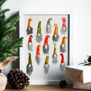 May include: Watercolor painting of colorful gnome figures in a white frame. The gnomes have various colored hats, including red, orange, and green, with gray beards. The artwork is displayed on a wooden surface with a pine cone and wrapped gift.