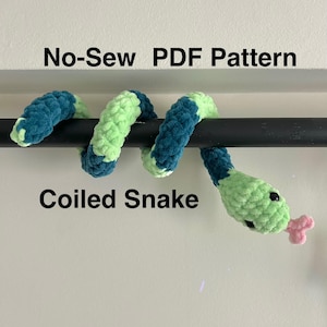May include: A green and blue coiled snake plush toy with a pink tongue. The toy is hanging from a black rod. The text "No-Sew PDF Pattern" and "Coiled Snake" are visible in the image.