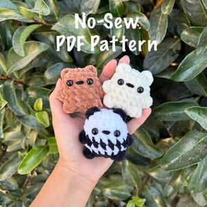 Op de afbeelding: Drie gehaakte berenvormige speeltjes in een hand, met de tekst "No-Sew PDF Pattern". Eén beer is bruin, één is wit en één is zwart en wit. Elk heeft zwarte knoopogen. De achtergrond is groen gebladerte.