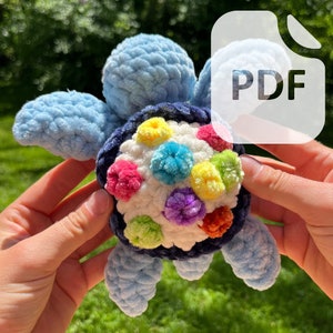 Op de afbeelding: Een gehaakte blauwe en witte schildpad met een kleurrijk bloemenontwerp op zijn schild. De schildpad is gemaakt van zacht garen en heeft een speelse, handgemaakte uitstraling.