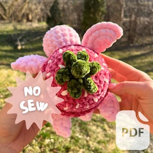 Puede incluir: Una fresa de crochet rosa y verde con detalles blancos. La fresa tiene un tallo y hojas de crochet verde. El texto "NO SEW" está en una forma de estrella rosa. El texto "PDF" está en un rectángulo blanco.