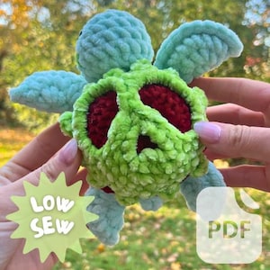 Puede incluir: Una tortuga de peluche tejida a crochet con una concha verde y un símbolo de paz rojo. La tortuga tiene aletas azules y una cabeza azul claro.
