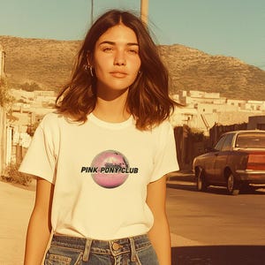 May include: A woman wearing a white t-shirt with a pink disco ball graphic and the text "PINK PONY CLUB". She is wearing blue jeans and has brown hair.