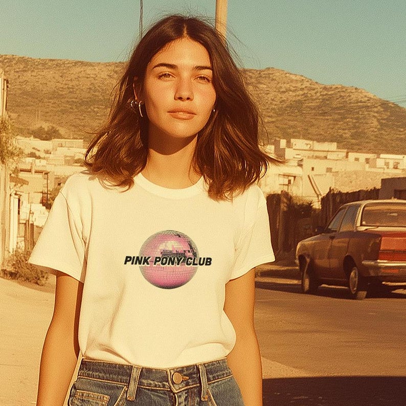 May include: A woman wearing a white t-shirt with a pink disco ball graphic and the text "PINK PONY CLUB". She is wearing blue jeans and has brown hair.