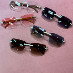 May include: Five pairs of rimless sunglasses with various tinted lenses, including clear, pink, and brown. The frames have a brown bridge and temples, with decorative silver accents. The sunglasses are arranged on a pink surface.